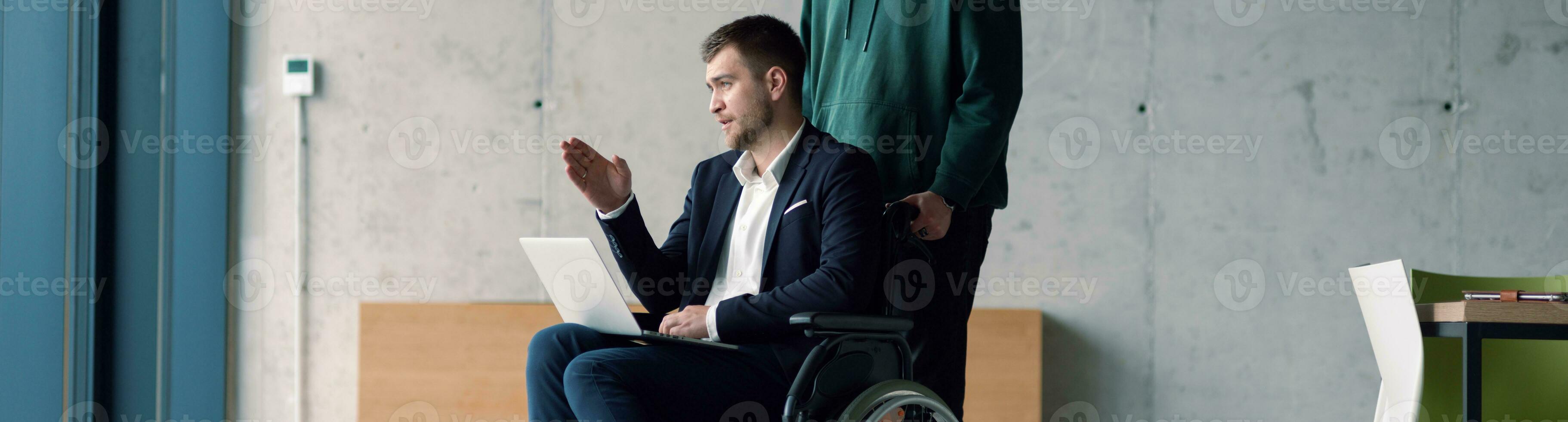 amplio cosecha de empresario en un silla de ruedas en un de moda oficina utilizando un ordenador portátil mientras detrás él es su negocio colega quien da él apoyo foto