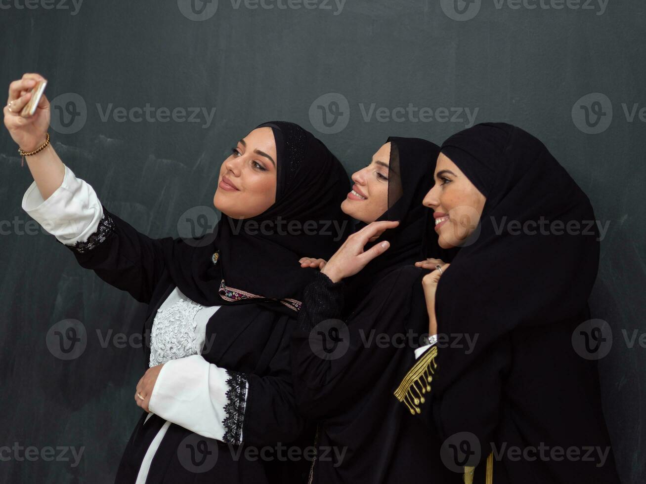 Group of young beautiful muslim women in fashionable dress with hijab using smartphone while taking selfie picture in front of black background photo