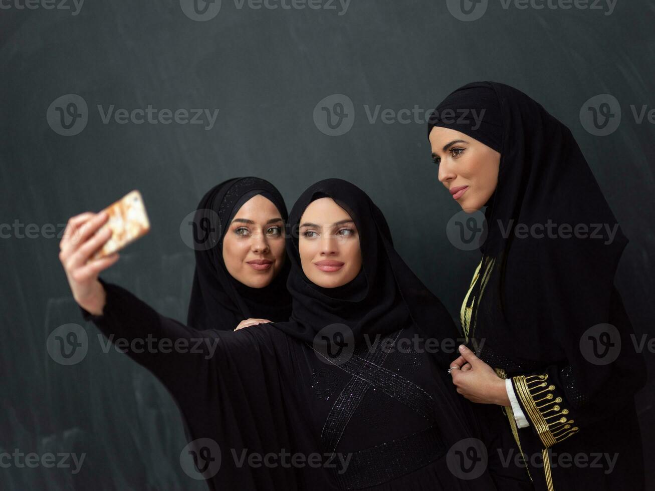Group of young beautiful muslim women in fashionable dress with hijab using smartphone while taking selfie picture in front of black background photo