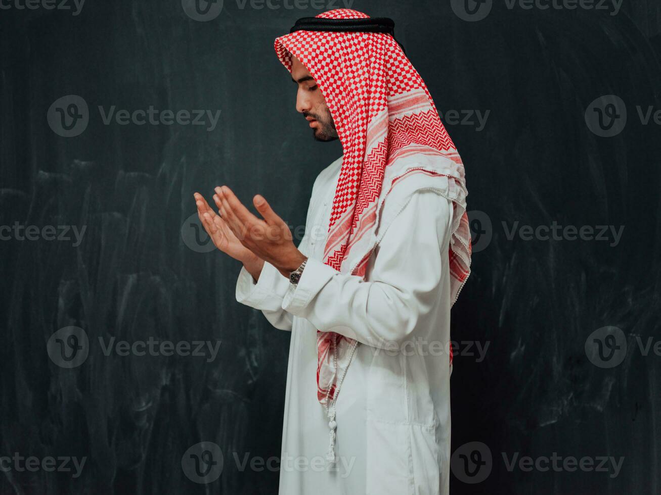 Arabian man in traditional clothes making traditional prayer to God, keeps hands in praying gesture in front of black chalkboard representing modern islam fashion and ramadan kareem concept photo