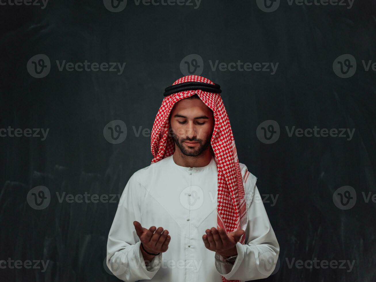Arabian man in traditional clothes making traditional prayer to God, keeps hands in praying gesture in front of black chalkboard representing modern islam fashion and ramadan kareem concept photo
