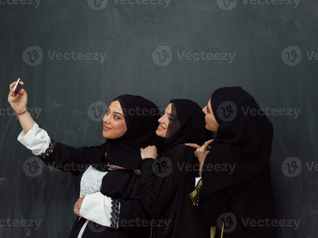 Group of young beautiful muslim women in fashionable dress with hijab using smartphone while taking selfie picture in front of black background photo