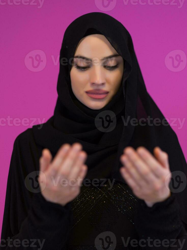 Muslim woman makes traditional prayer to God, keeps hands in praying gesture, wears traditional white clothes, has serious facial expression, isolated over plastic pink background photo