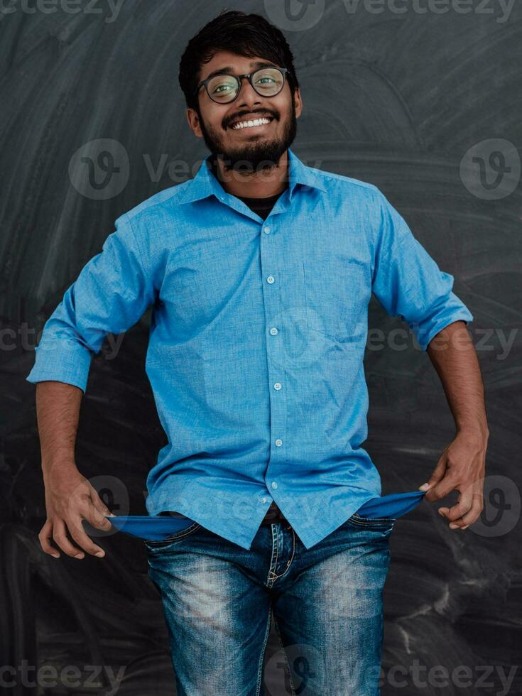 Photo of an Indian man wearing a blue shirt and glasses while symbolically showing his empty pockets on the background of a school blackboard.