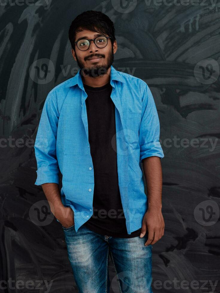 Indian smiling young student in blue shirt and glasses posing on school blackboard background photo