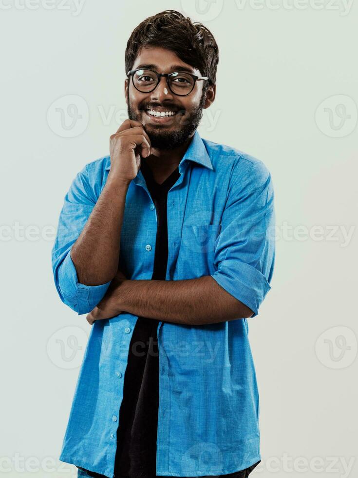 A young Indian man in a blue shirt and glasses poses thoughtfully in front of a gray background photo