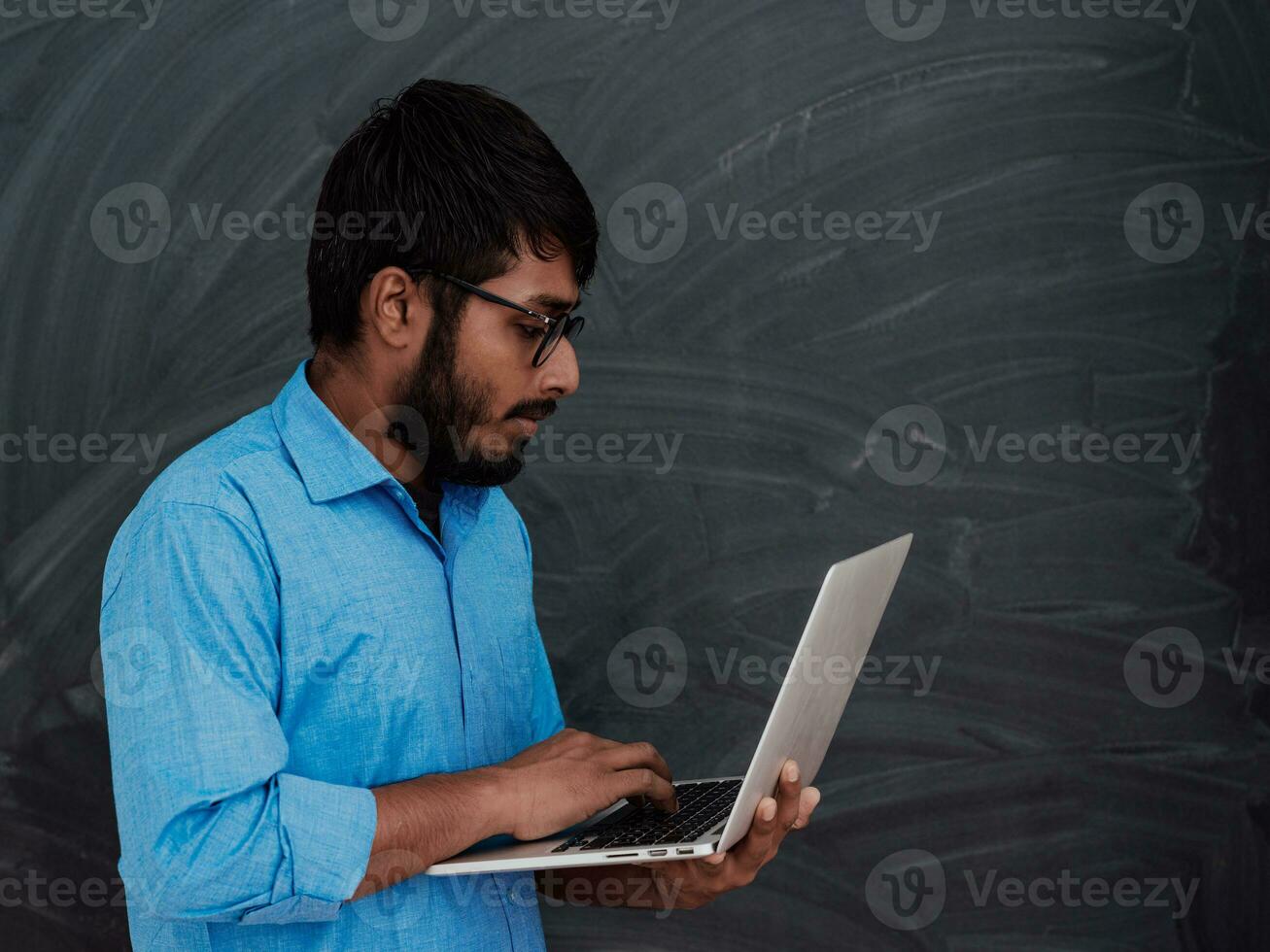 Indian smiling young student in blue shirt and glasses using laptop and posing on school blackboard background photo