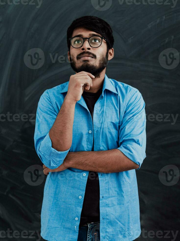 A young Indian man in a blue shirt and glasses poses thoughtfully in front of school board photo