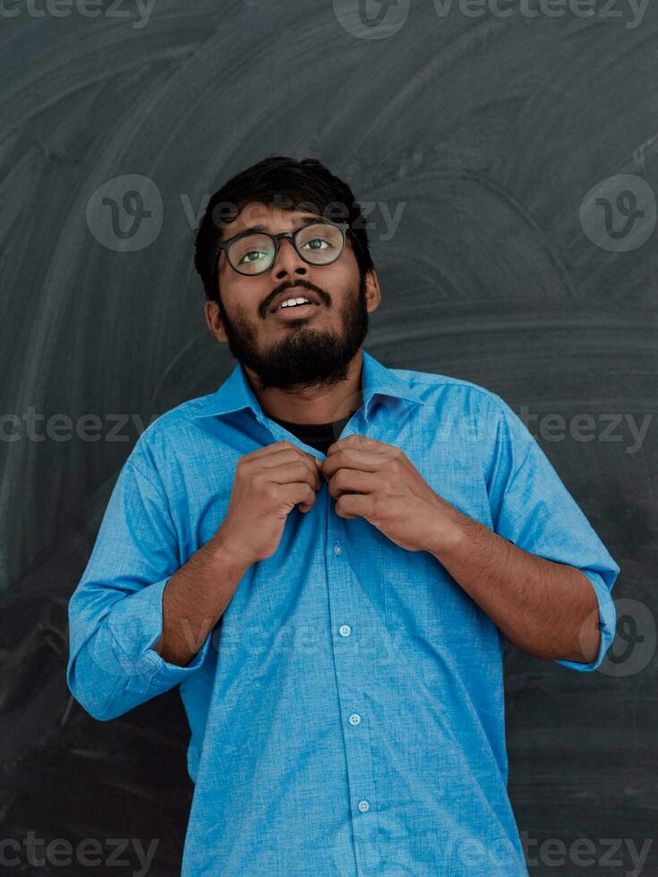 Indian young student wearing glasses puts on a blue shirt while standing in front of the school blackboard photo