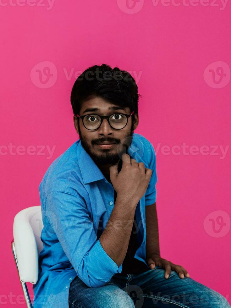 A young Indian student in a blue shirt with glasses sitting in front of a pink background photo