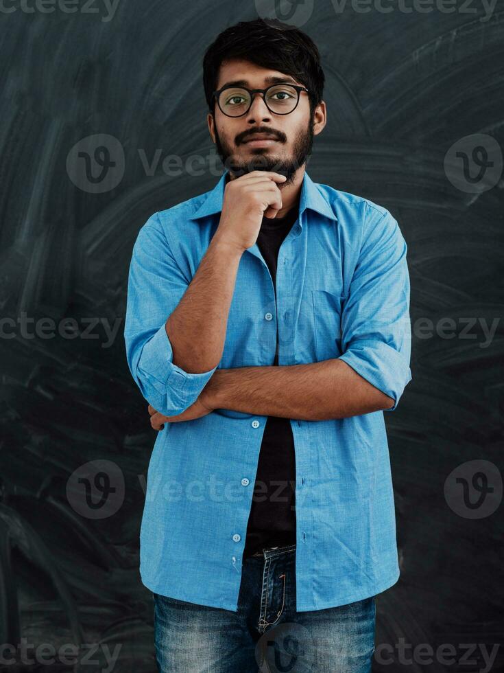 A young Indian man in a blue shirt and glasses poses thoughtfully in front of school board photo