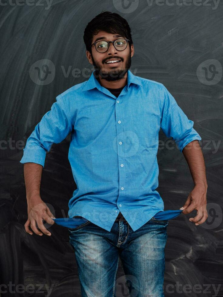Photo of an Indian man wearing a blue shirt and glasses while symbolically showing his empty pockets on the background of a school blackboard.