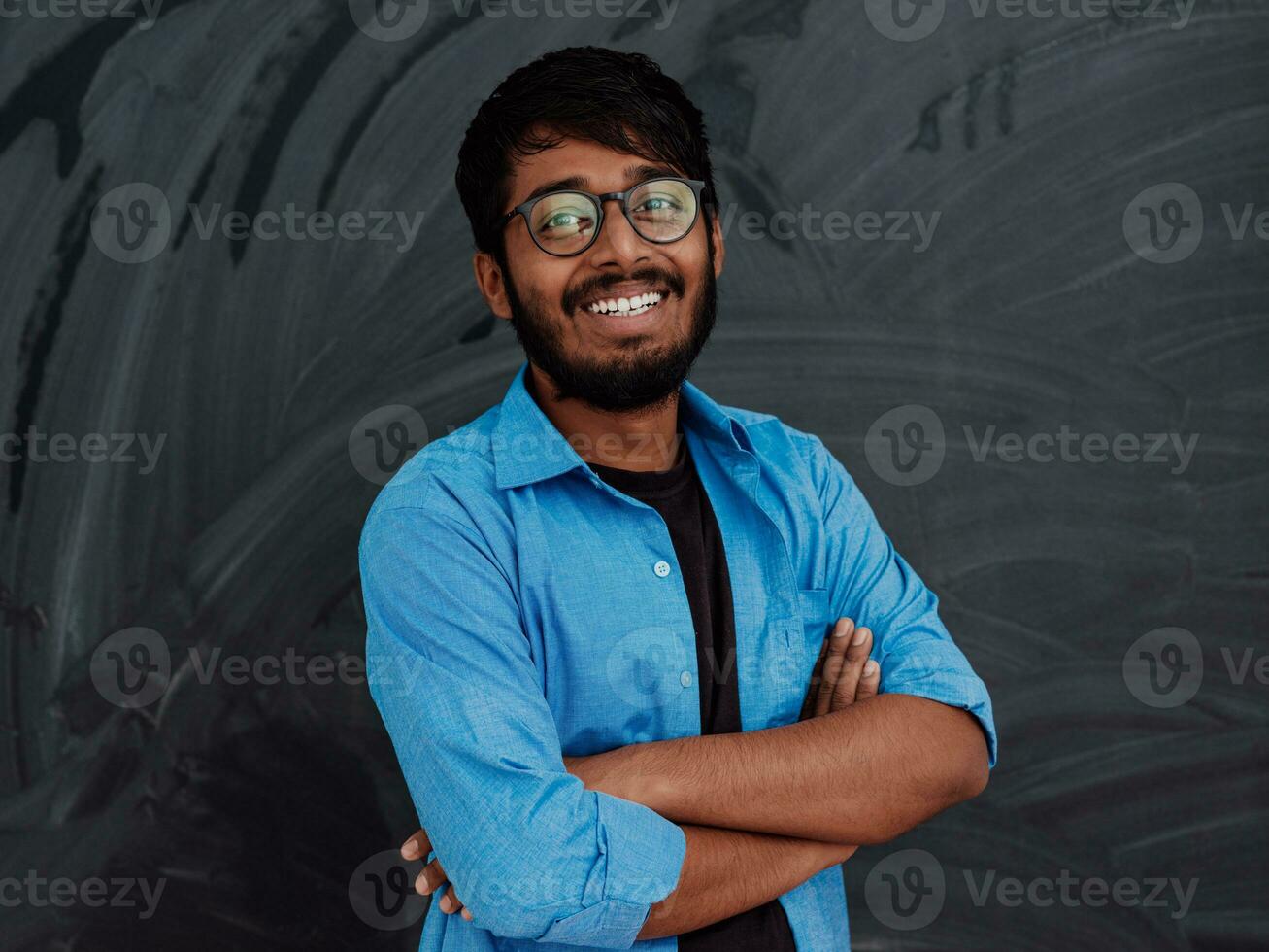 A young Indian student in a blue shirt with glasses posing with his arms crossed in front of the school blackboard photo