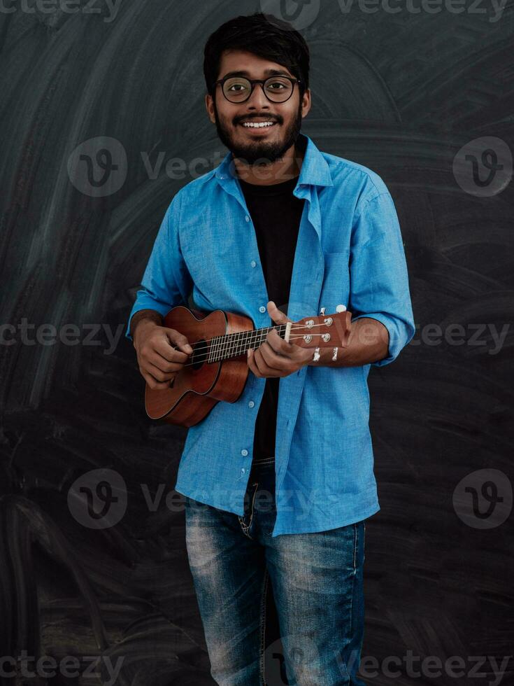 Indian young man in a blue shirt and glasses playing the guitar in front of the school blackboard photo