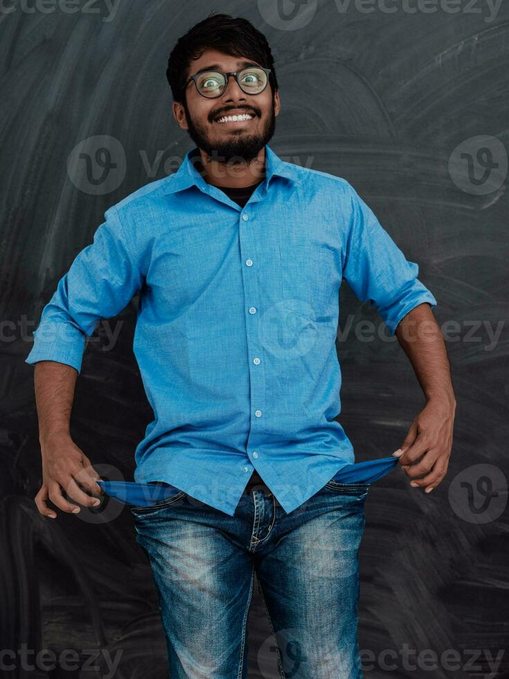 Photo of an Indian man wearing a blue shirt and glasses while symbolically showing his empty pockets on the background of a school blackboard.