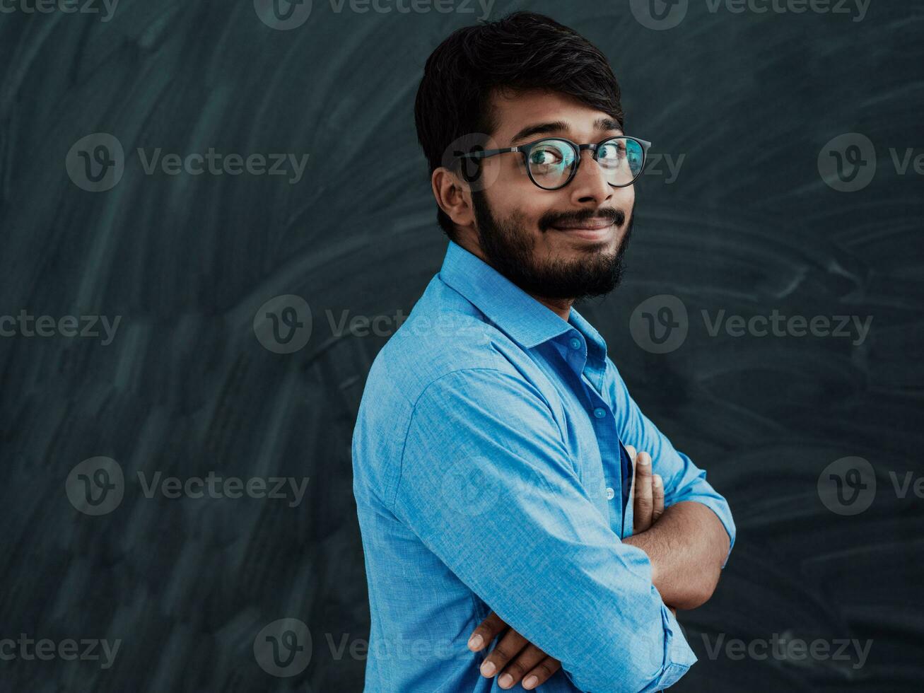 A young Indian student in a blue shirt with glasses posing with his arms crossed in front of the school blackboard photo