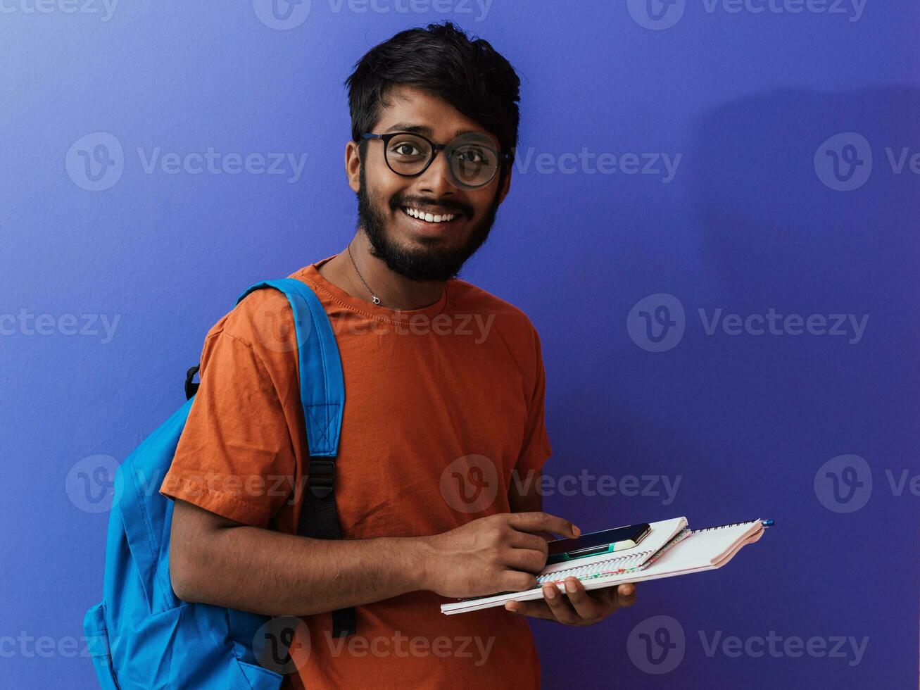 indio estudiante con azul mochila, lentes y cuaderno posando en púrpura antecedentes. el concepto de educación y enseñanza. hora a Vamos espalda a colegio foto