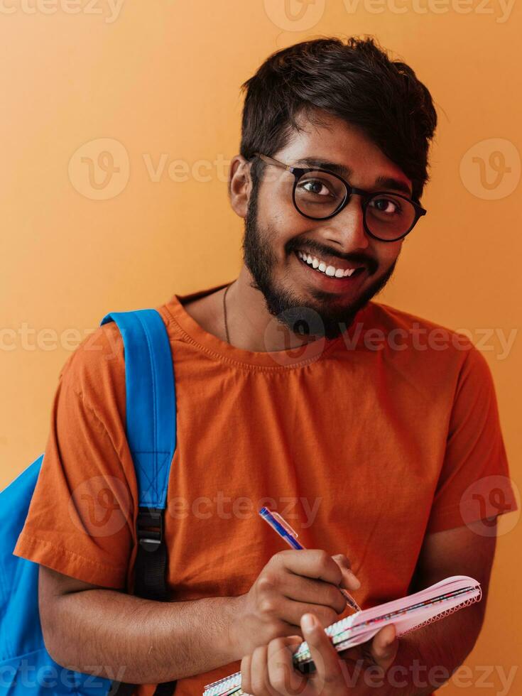 indio estudiante con azul mochila, lentes y cuaderno posando en naranja antecedentes. el concepto de educación y enseñanza. hora a Vamos espalda a colegio foto