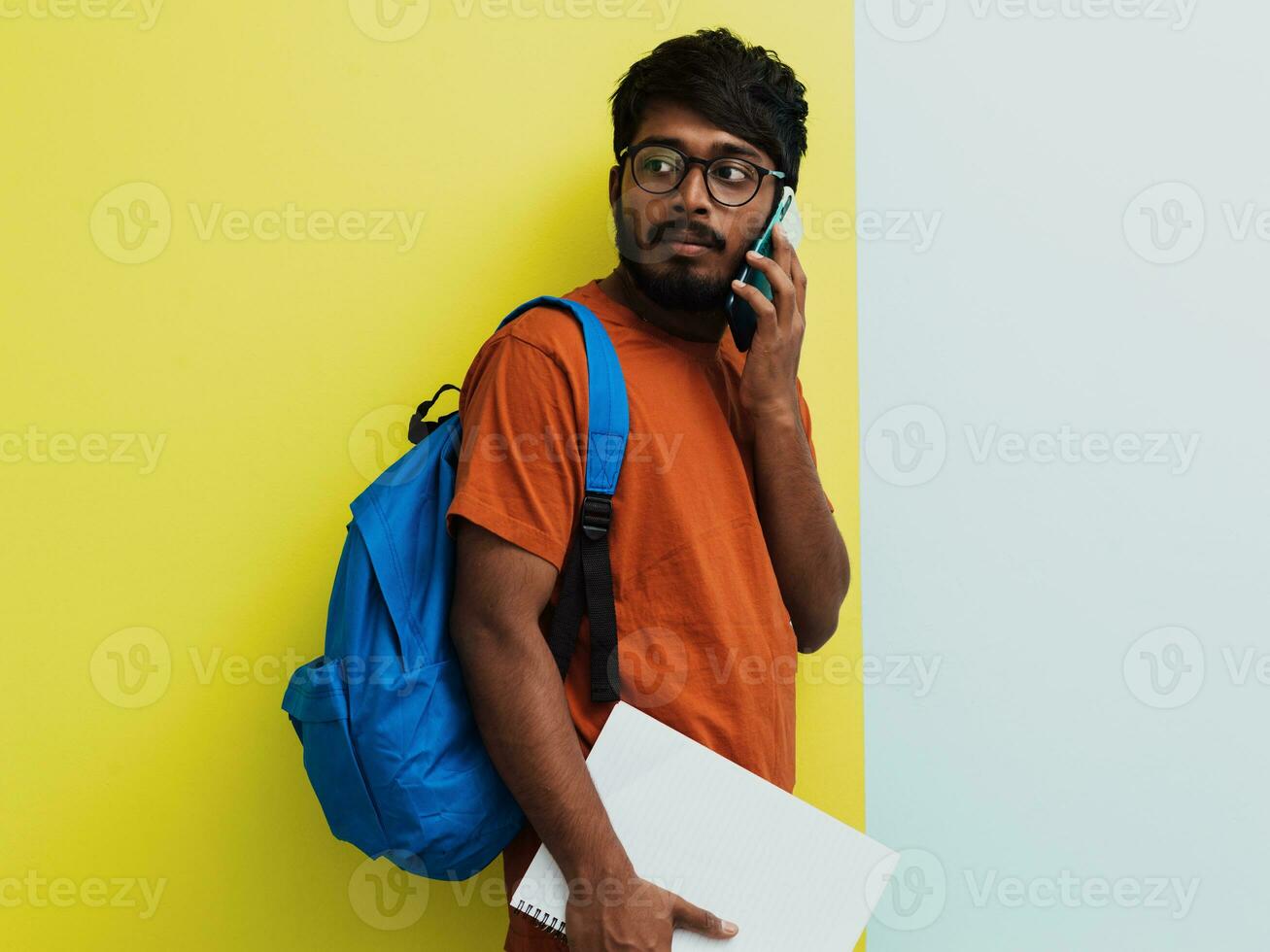 Indian student with blue backpack, glasses and notebook using smarpthone and posing on green-grey background. The concept of education and schooling. Time to go back to school photo