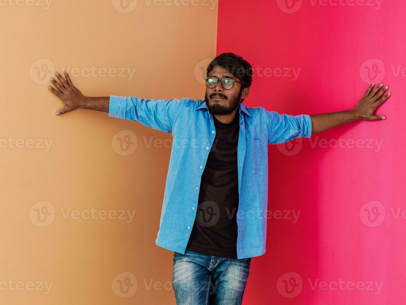 A young Indian student in a blue shirt with glasses posing on pink-orange background photo