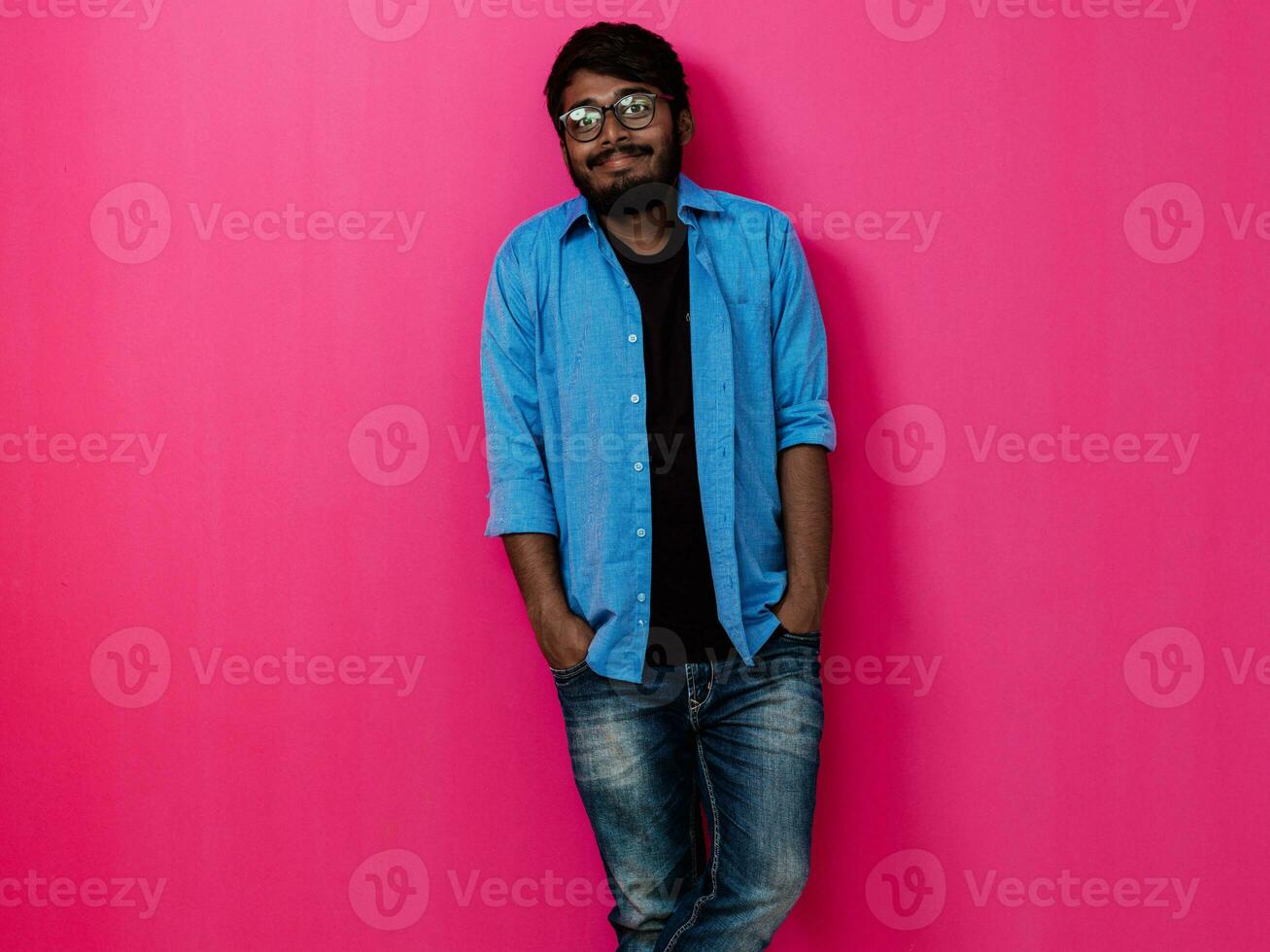 indio sonriente joven hombre con azul camisa y lentes posando en rosado antecedentes foto