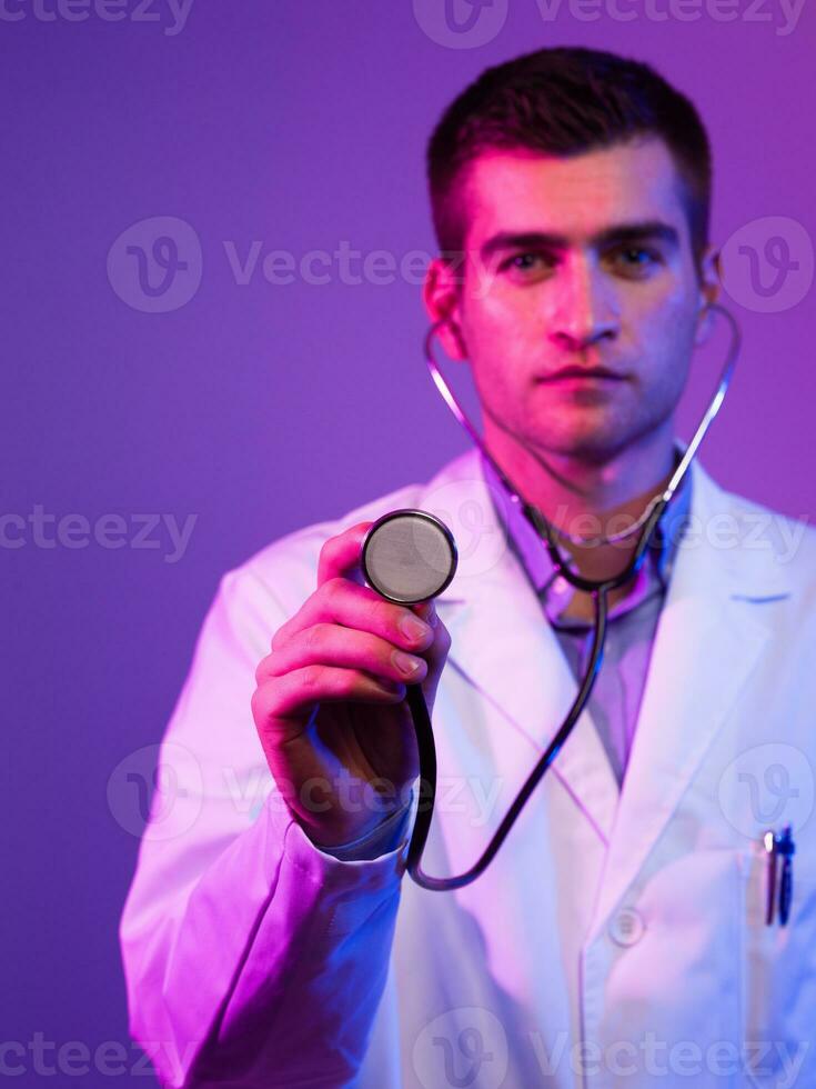 Portrait of hero in white coat. Cheerful smiling young doctor with stethoscope in medical hospital standing against blue and pink background. Coronavirus covid-19 danger alert photo