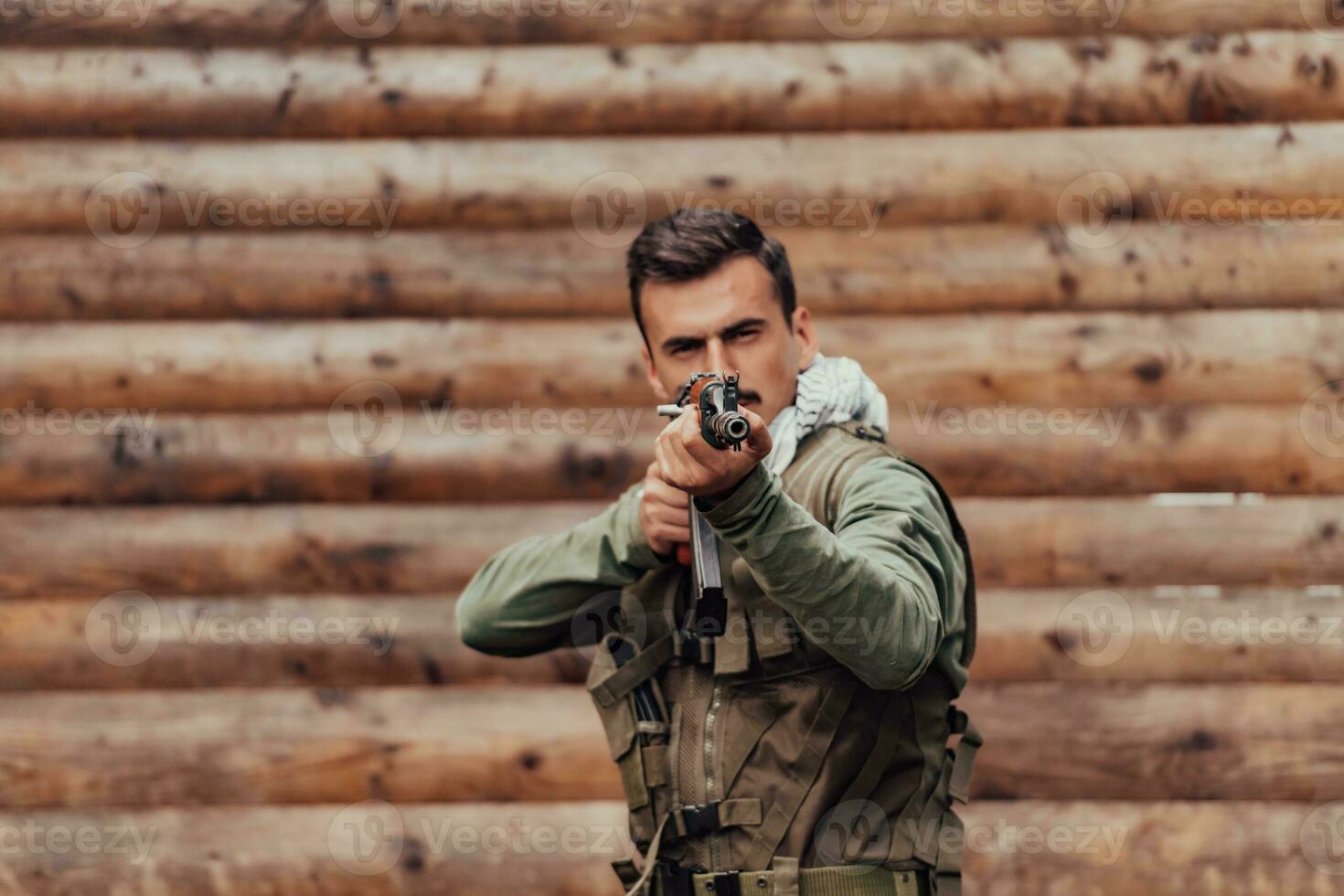 A soldier in uniform with a rifle in his hand is standing in front of a wooden wall. A soldier guards the forest base from the enemy photo