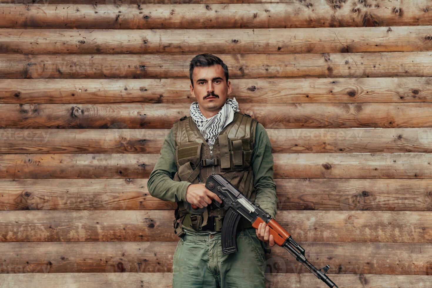 A soldier in uniform with a rifle in his hand is standing in front of a wooden wall. A soldier guards the forest base from the enemy photo