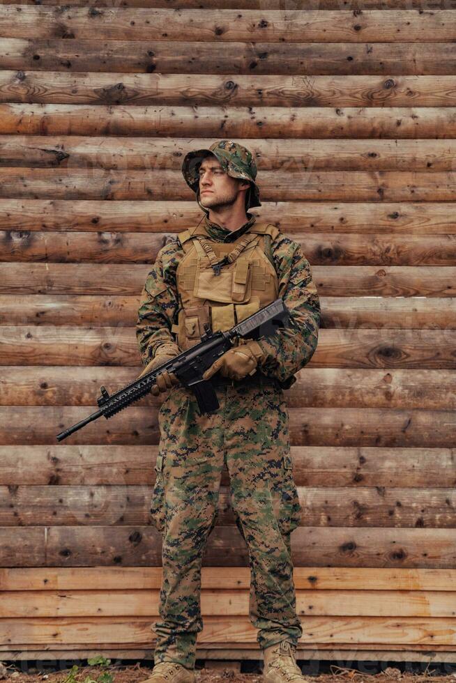 A soldier in uniform with a rifle in his hand is standing in front of a wooden wall. A soldier guards the forest base from the enemy photo