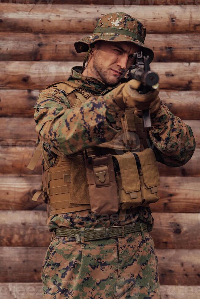 A soldier in uniform with a rifle in his hand is standing in front of a wooden wall. A soldier guards the forest base from the enemy photo