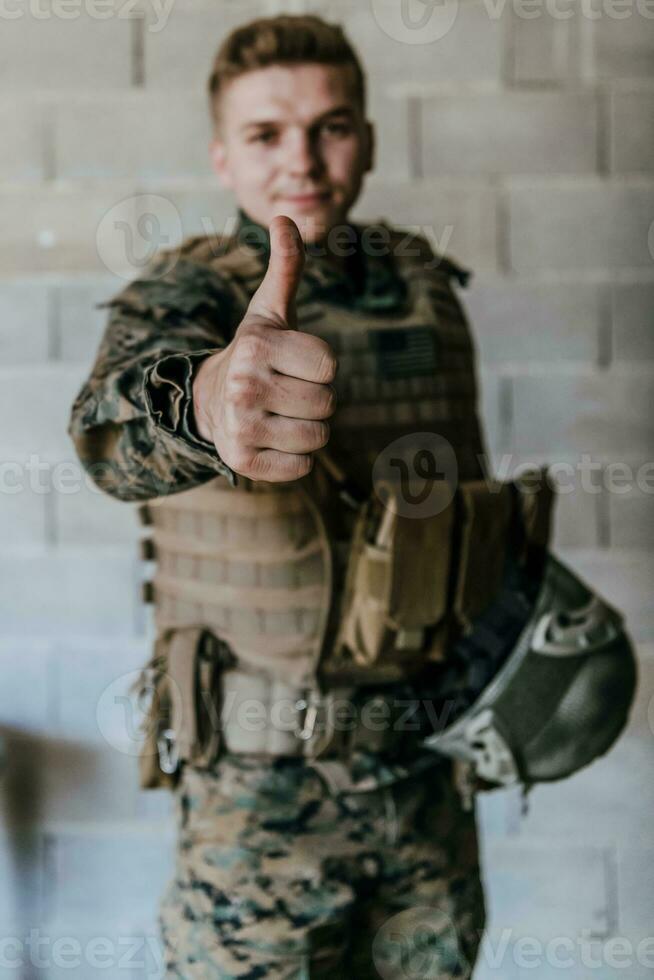 The soldier makes a gesture of success with his hand. A soldier in full war gear stands in front of a stone wall and shows the ok sign with his finger photo