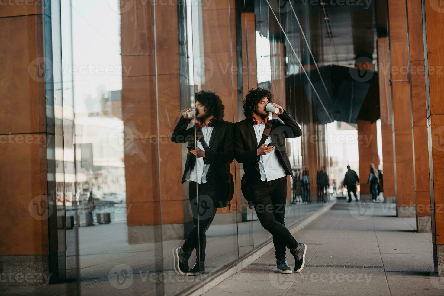 Trendy businessman with leather briefcase in financial district using smartphone while listening audio songs headphones drinking coffee photo