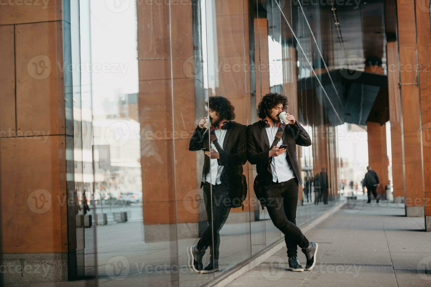 Trendy businessman with leather briefcase in financial district using smartphone while listening audio songs headphones drinking coffee photo