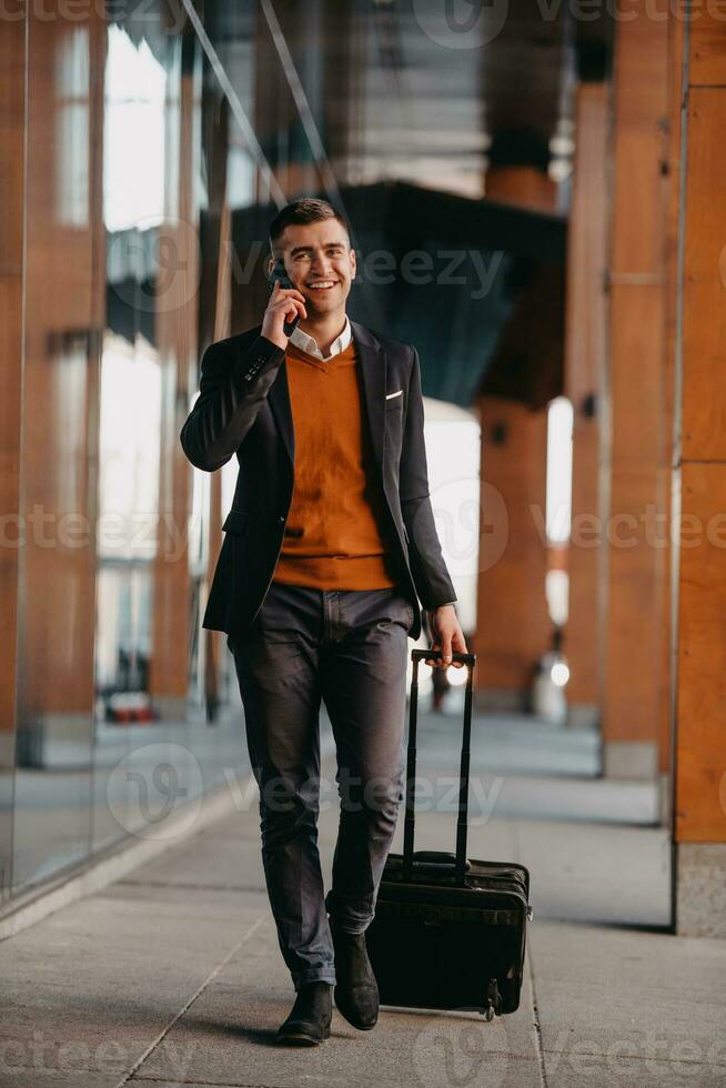 Going to airport terminal. Confident businessman traveler walking on city streets and pulling his suitcase drinking coffee and speaking on smartphone photo