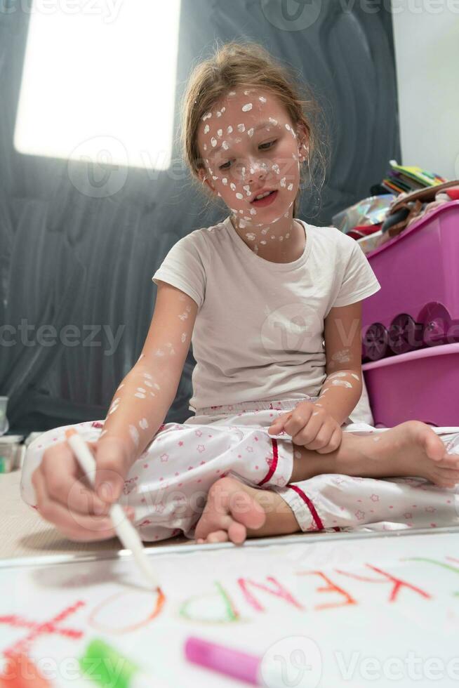 pequeño colegio niña con varicela dibujo en blanco tablero en niños' habitación, antiséptico crema aplicado a cara y cuerpo. pizarra y juguetes antecedentes. foto