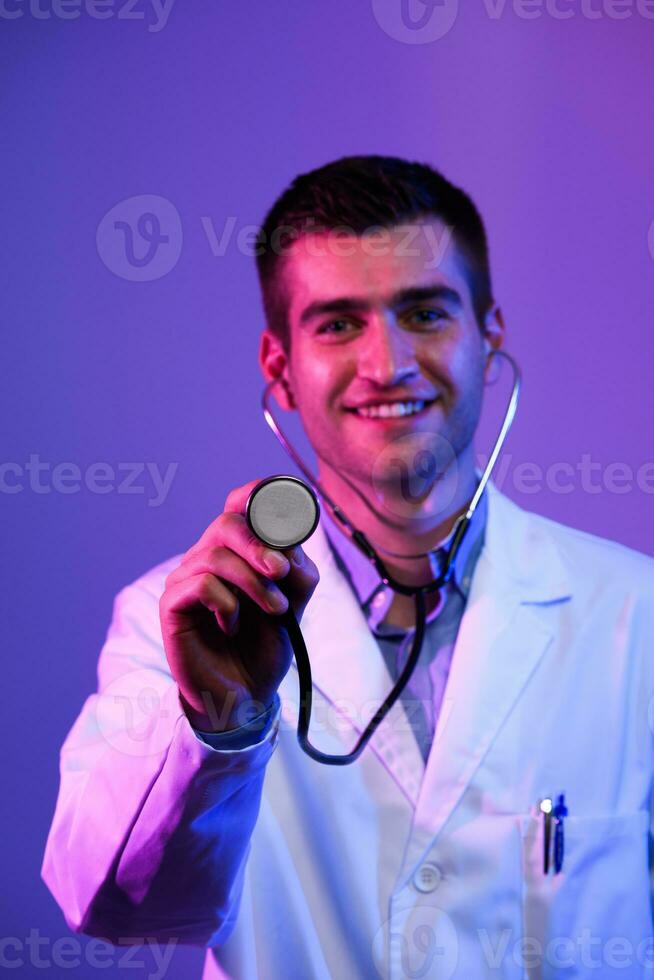 Portrait of hero in white coat. Cheerful smiling young doctor with stethoscope in medical hospital standing against blue and pink background. Coronavirus covid-19 danger alert photo