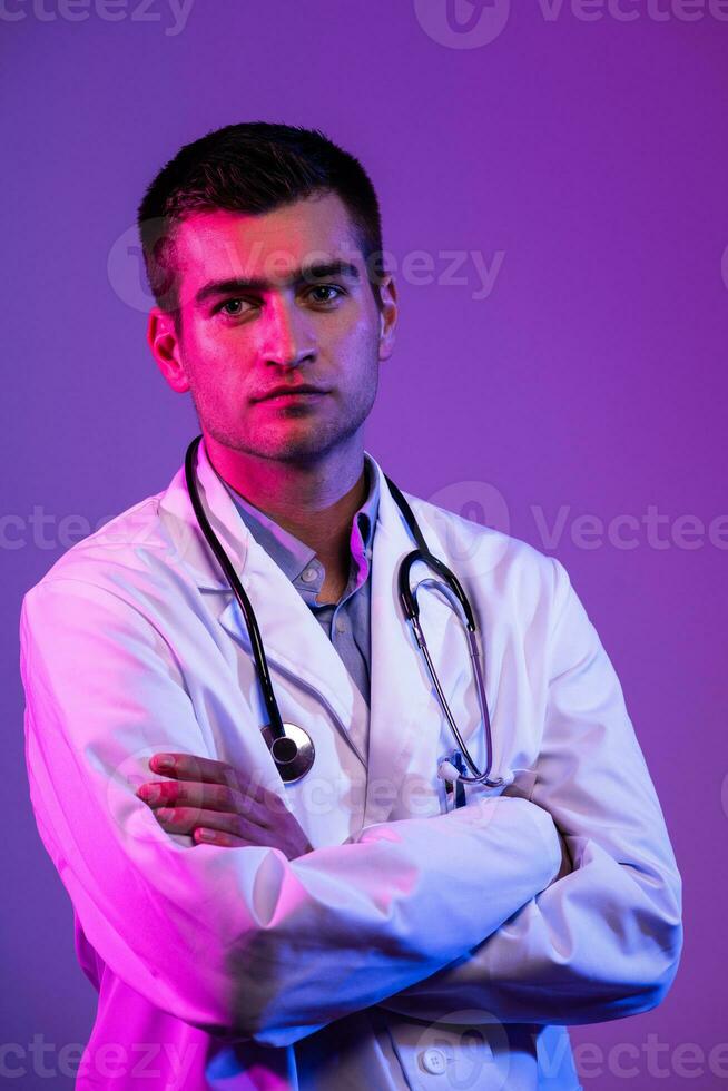 Portrait of hero in white coat. Cheerful smiling young doctor with stethoscope in medical hospital standing against blue and pink background. Coronavirus covid-19 danger alert photo