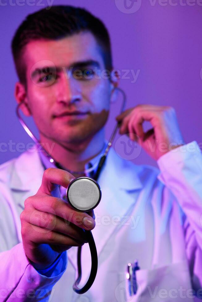 Portrait of hero in white coat. Cheerful smiling young doctor with stethoscope in medical hospital standing against blue and pink background. Coronavirus covid-19 danger alert photo