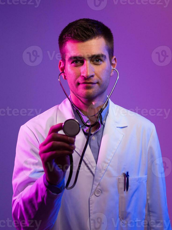 Portrait of hero in white coat. Cheerful smiling young doctor with stethoscope in medical hospital standing against blue and pink background. Coronavirus covid-19 danger alert photo
