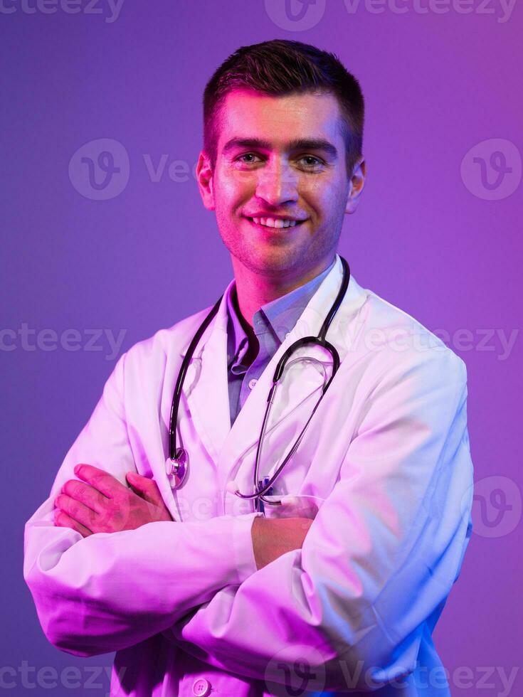 Portrait of hero in white coat. Cheerful smiling young doctor with stethoscope in medical hospital standing against blue and pink background. Coronavirus covid-19 danger alert photo