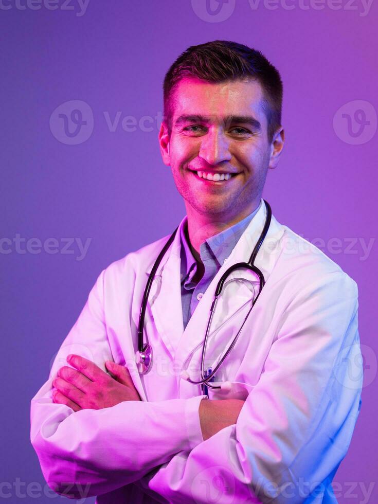 Portrait of hero in white coat. Cheerful smiling young doctor with stethoscope in medical hospital standing against blue and pink background. Coronavirus covid-19 danger alert photo
