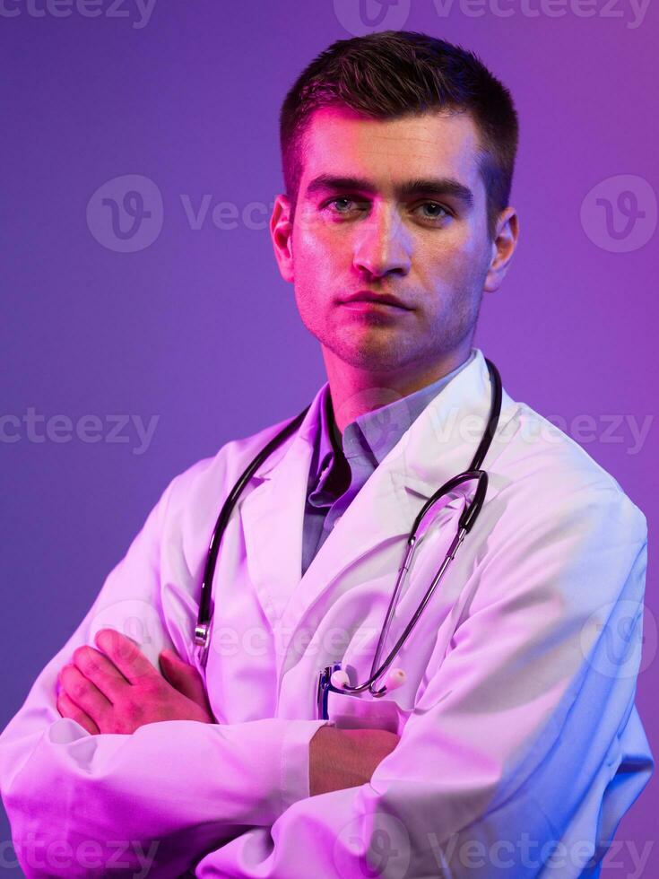 Portrait of hero in white coat. Cheerful smiling young doctor with stethoscope in medical hospital standing against blue and pink background. Coronavirus covid-19 danger alert photo