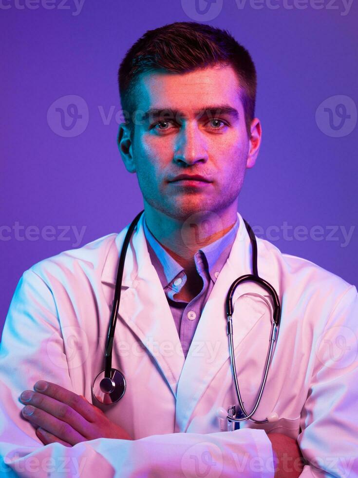 Portrait of hero in white coat. Cheerful smiling young doctor with stethoscope in medical hospital standing against blue and pink background. Coronavirus covid-19 danger alert photo