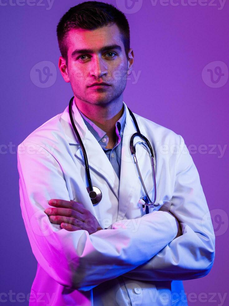 Portrait of hero in white coat. Cheerful smiling young doctor with stethoscope in medical hospital standing against blue and pink background. Coronavirus covid-19 danger alert photo