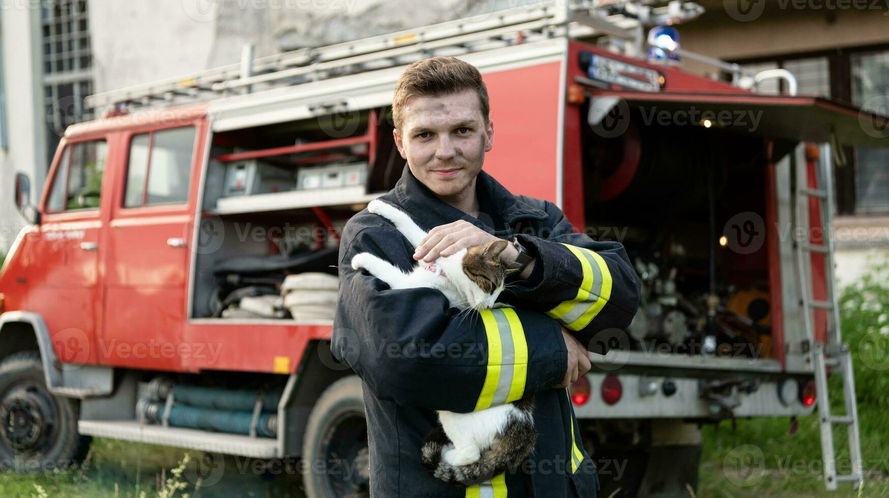 Close-up portrait of heroic fireman in protective suit and red helmet holds saved cat in his arms. Firefighter in fire fighting operation. photo