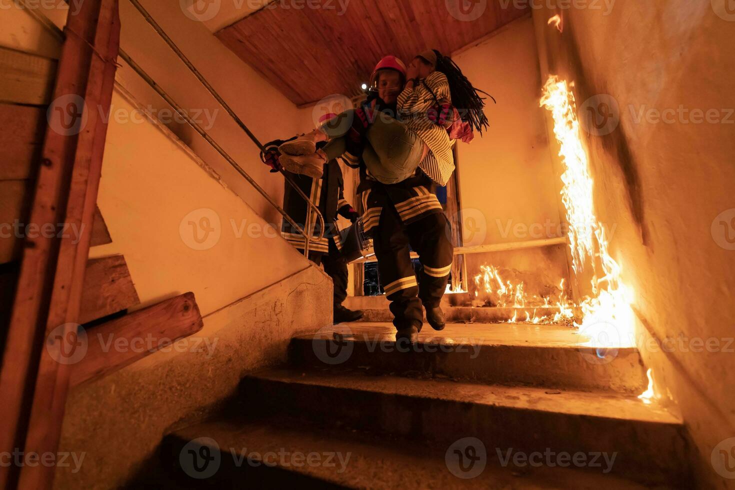 Brave Fireman Descends Stairs of a Burning Building and Holds Saved Girl in His Arms. Open fire and one Firefighter in the Background. photo