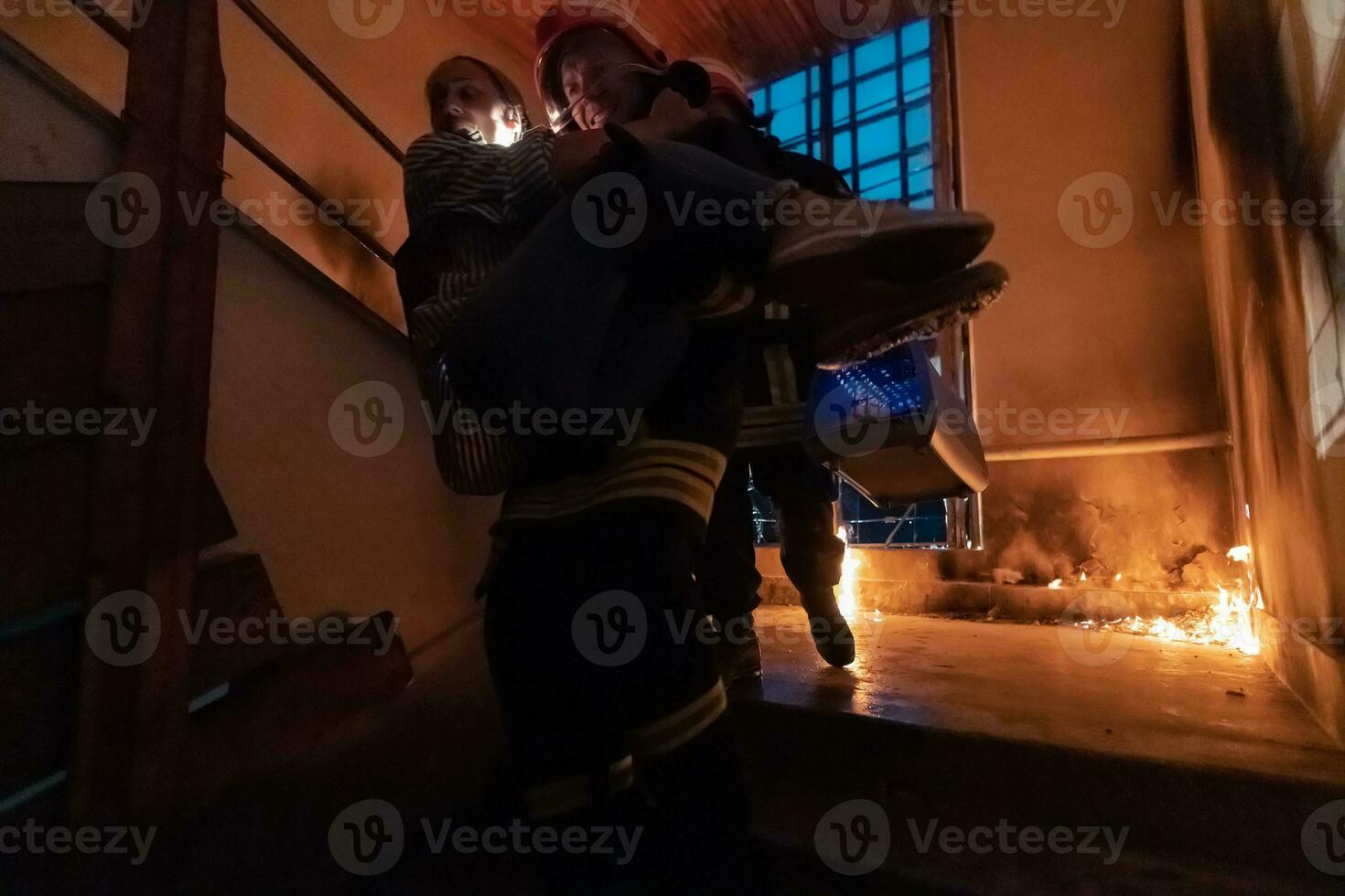 Brave Fireman Descends Stairs of a Burning Building and Holds Saved Girl in His Arms. Open fire and one Firefighter in the Background. photo