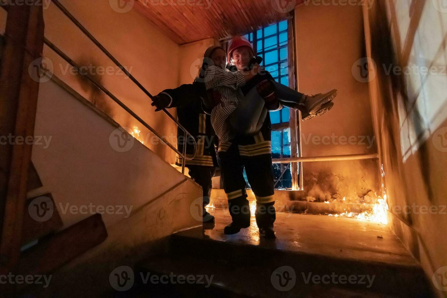 Brave Fireman Descends Stairs of a Burning Building and Holds Saved Girl in His Arms. Open fire and one Firefighter in the Background. photo