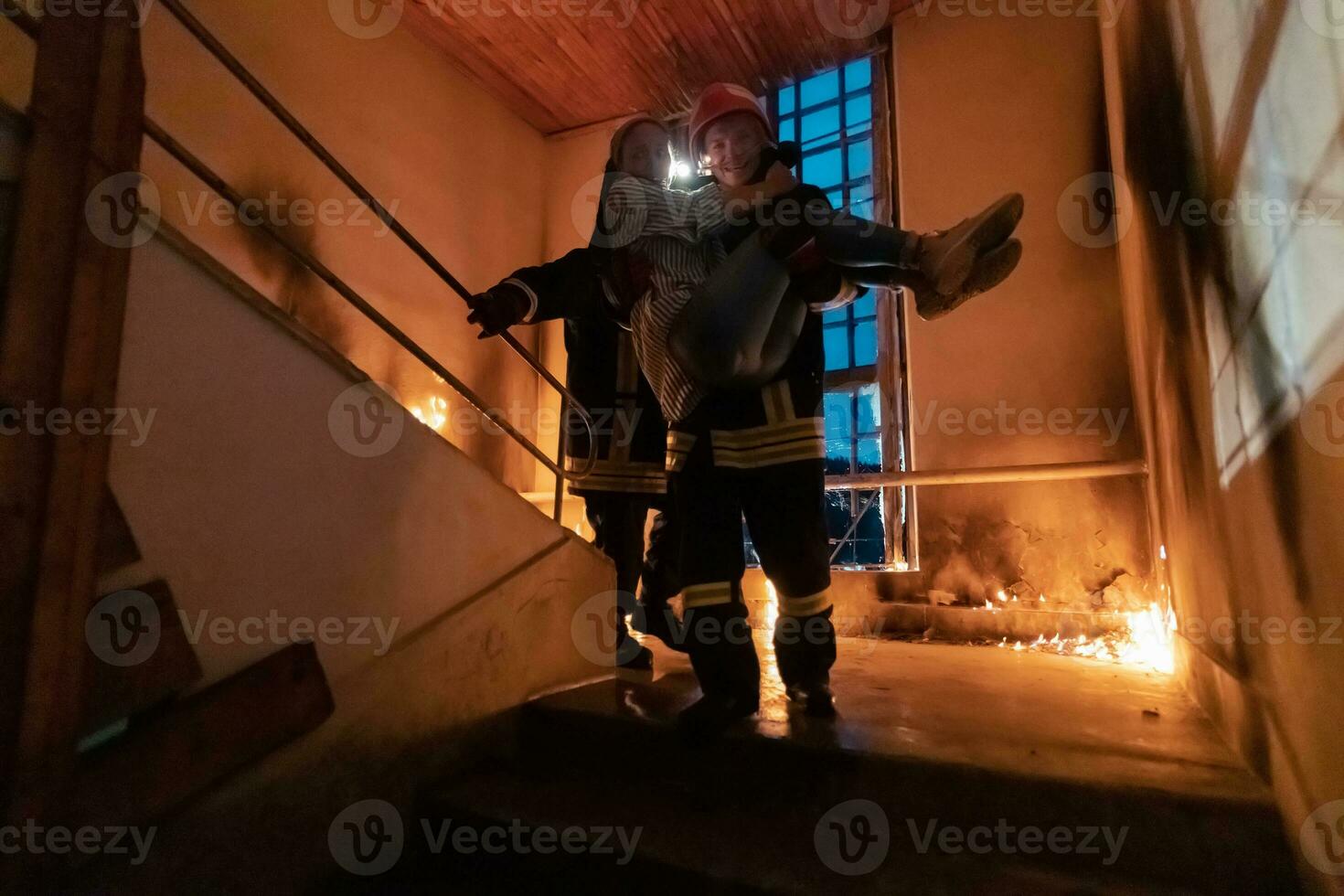 Brave Fireman Descends Stairs of a Burning Building and Holds Saved Girl in His Arms. Open fire and one Firefighter in the Background. photo