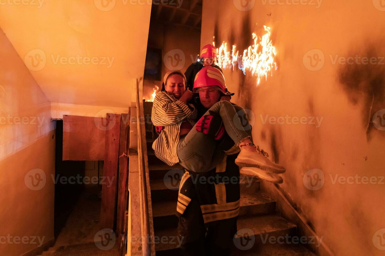 Brave Fireman Descends Stairs of a Burning Building and Holds Saved Girl in His Arms. Open fire and one Firefighter in the Background. photo